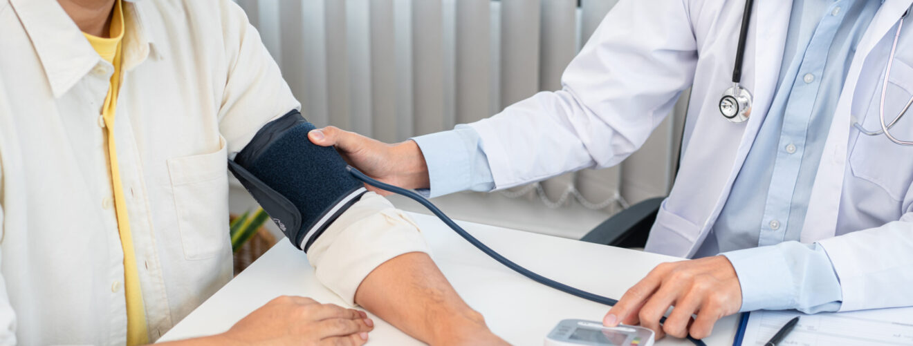 Male doctor in medical face mask is using sphygmomanometer to checking blood pressure to male patient and taking note data on clipboard in clinic
