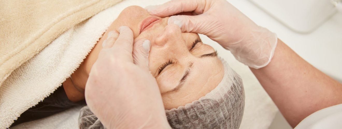 Charming woman lying on daybed and smiling while receiving rejuvenation facial massage at wellness center